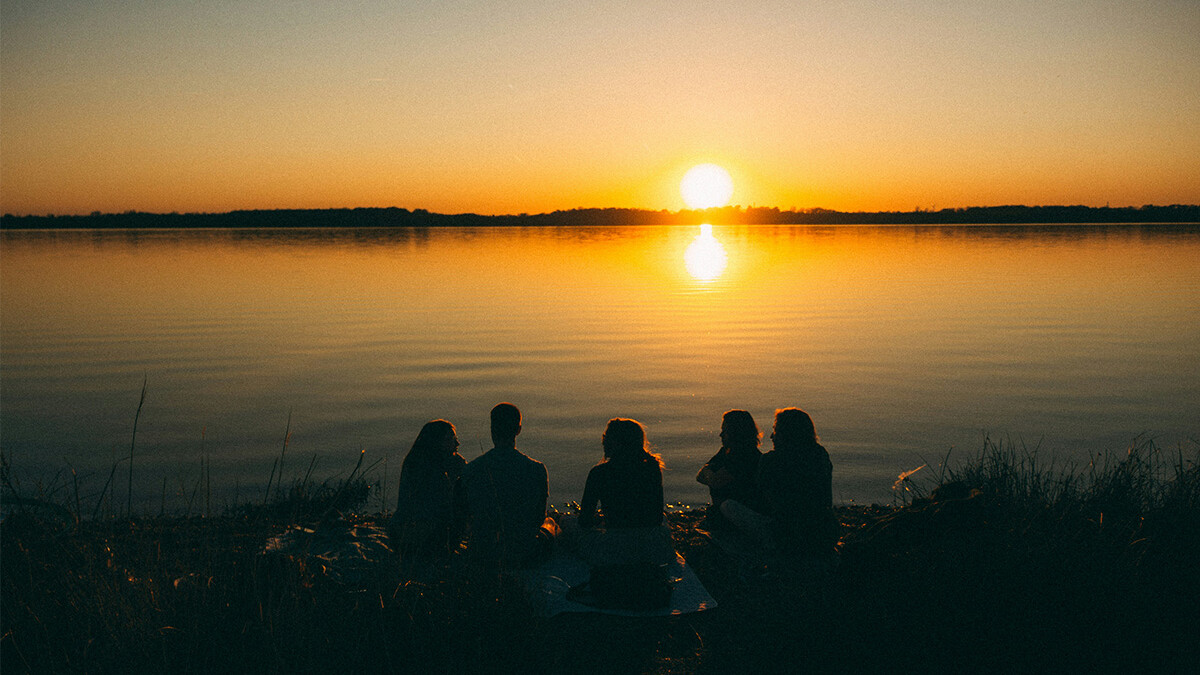 Gruppe junger Menschen sitzt am Ufer vom See und schaut Sonnenuntergang