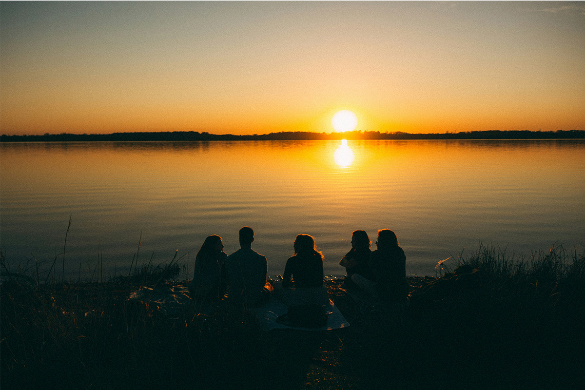 Gruppe junger Menschen sitzt am Ufer vom See und schaut Sonnenuntergang